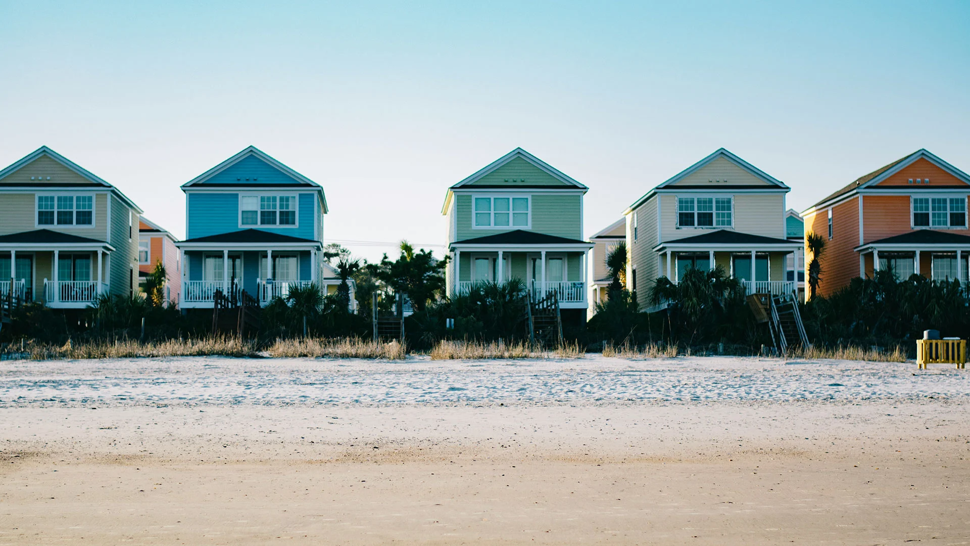 A row of colorful two-story beach houses behind a sandy dune on a clear summer day.